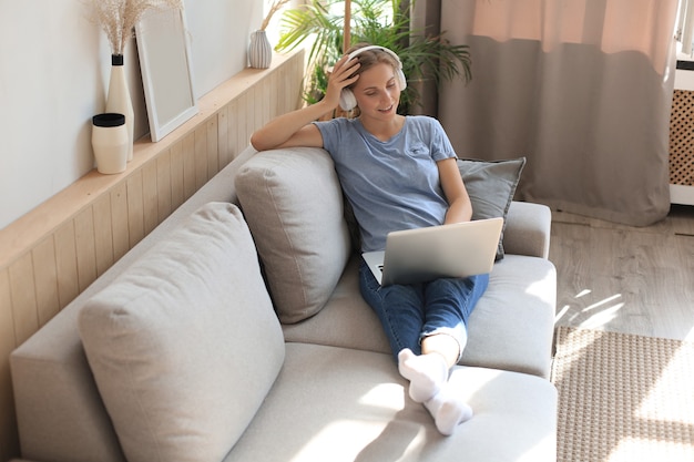 Sorridente giovane donna con cuffie e laptop sul divano.