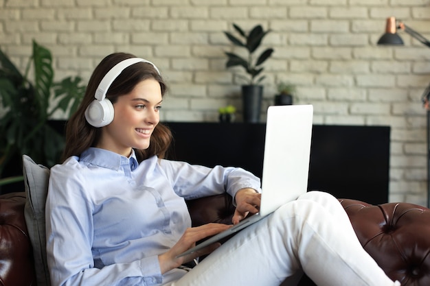 Smiling young woman with headphones and laptop on the sofa.