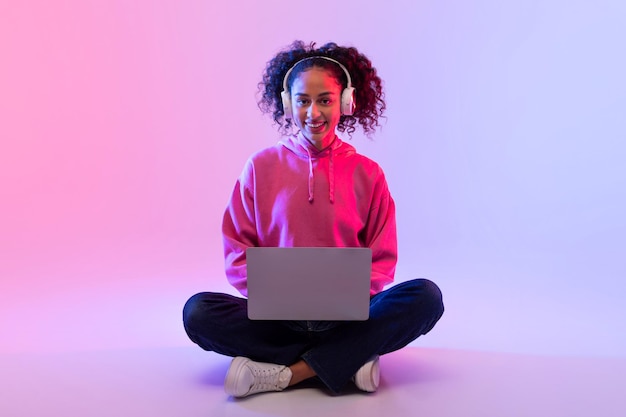 Smiling young woman with laptop and headphones sitting on the floor with gradient background