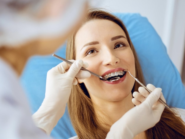 歯科医院で歯科医によって検査された歯列矯正ブラケットを持つ笑顔の若い女性。健康な歯と薬の概念。
