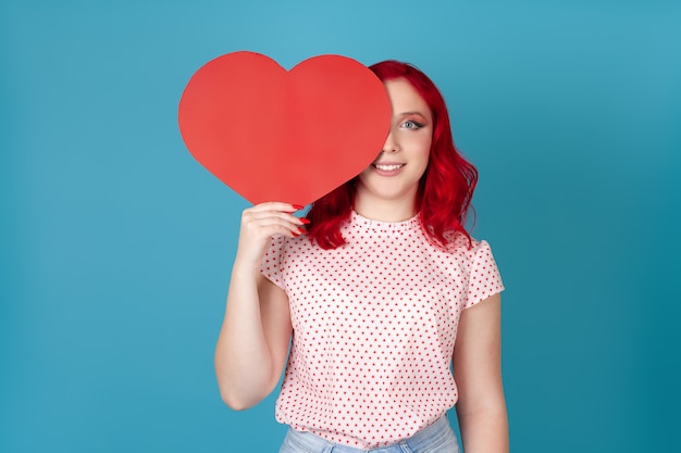 Улыбающаяся молодая женщина с рыжими волосами прячет половину лица за красным бумажным сердцем