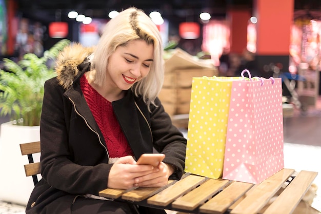 Фото Улыбающаяся молодая женщина с сумками для покупок использует телефон в ресторане