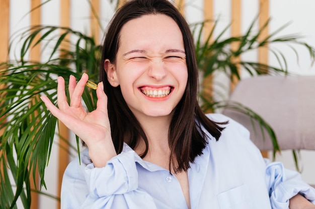 Smiling young woman with short hair holding pill omega capsule in hand at home Girl taking medicine indoors