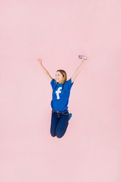 Giovane donna sorridente con lo smartphone che salta sul fondo rosa