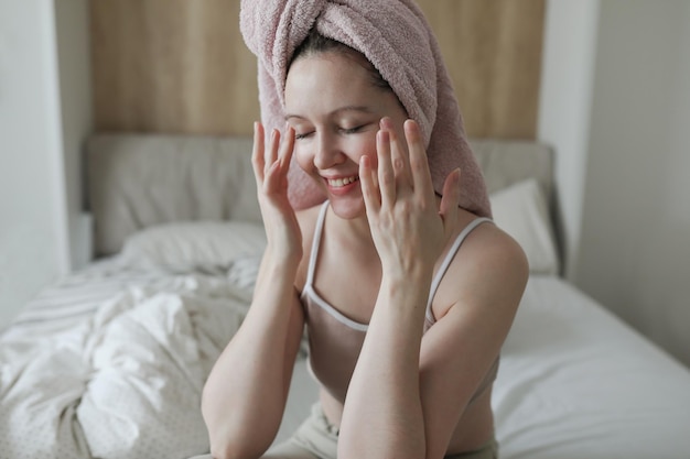 Smiling young woman with a towel on head apply cream on face skin