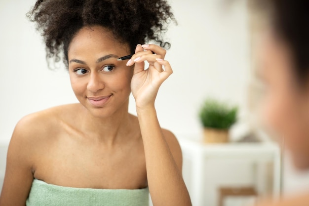 Smiling young woman with tweezers plucking her eyebrow and looking in mirror at home bathroom