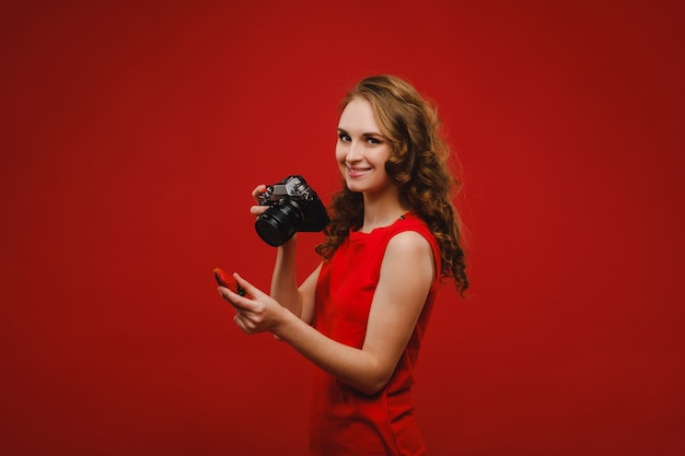 A smiling young woman with wavy hair holds a strawberry and photographs it, holding a delicious fresh strawberry on a bright red background.