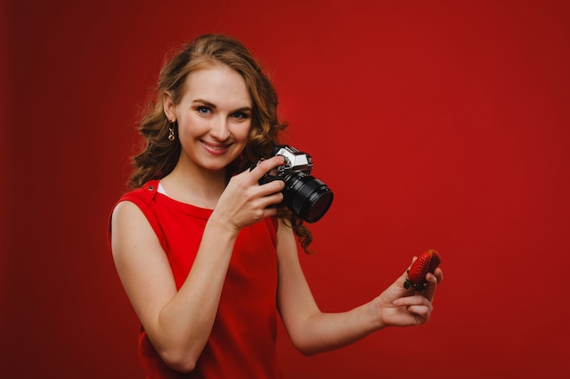 A smiling young woman with wavy hair holds a strawberry and photographs it, holding a delicious fresh strawberry on a bright red background