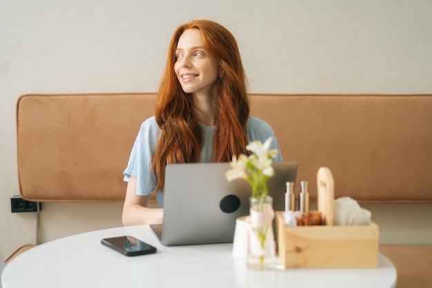 居心地の良いカフェのテーブルに座って、目をそらして、ラップトップコンピューターを使用して作業している笑顔の若い女性。