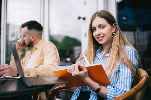 Smiling young woman writing on notebook sitting with man having phone call in cafe