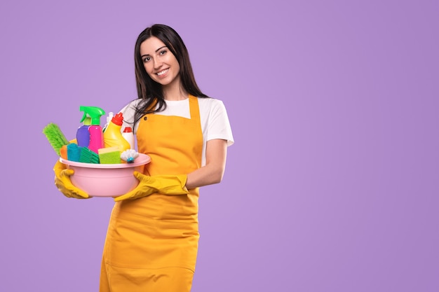 Smiling young woman in yellow apron and gloves carrying basin with colorful bottles with detergents and tools for house cleaning against violet background