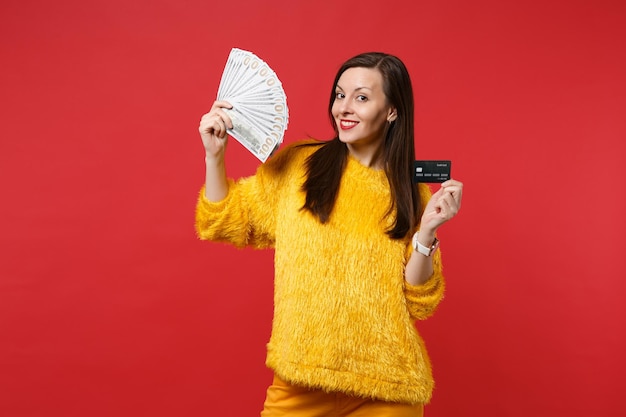 Smiling young woman in yellow fur sweater hold credit bank card, fan of money in dollar banknotes, cash money isolated on red background. People sincere emotions lifestyle concept. Mock up copy space.