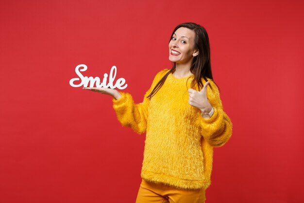 Улыбающаяся молодая женщина в желтом меховом свитере показывает палец вверх, держа деревянную улыбку букв слова, изолированную на фоне красной стены в студии. Люди искренние эмоции, концепция образа жизни. Копируйте пространство для копирования.