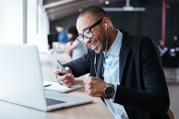 Photo smilling cheerful businessman texting message on the smartphone in front of the laptop in the office