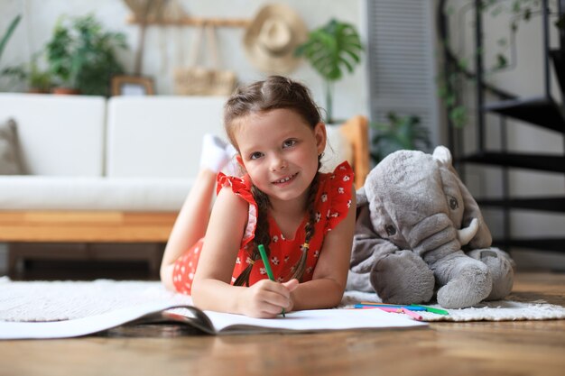 Smilling happy girl lying on warm floor with a toy elephant near to her enjoying creative activity, drawing pencils coloring pictures in albums.