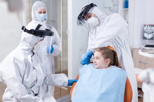 Улыбающаяся девочка на стоматологическом осмотре в комбинезоне из-за пандемии коронавируса. Стоматолог во время covid19 в костюме ppe делает процедуру зубов ребенка, сидящего на ch