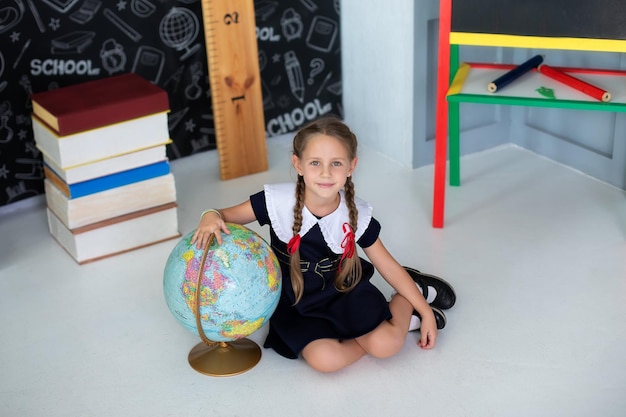 Фото Улыбающаяся маленькая девочка в школьной форме и с косичками держит глобус в классе.