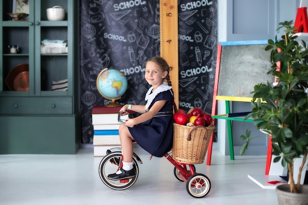 Smilling little girl in a school uniform on red tricycle with basket apples in classroom