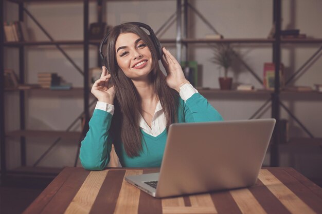 Smilling woman with headphones is sitting at the wood table with grey laptop