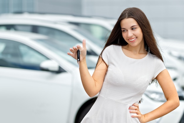 Smilling woman with keys and agreement on background of car dealership