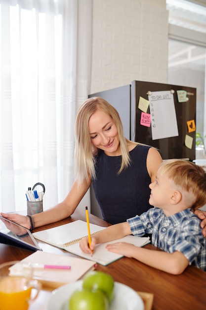 Smilng young mother with digital tablet looking at her son doing homework and writing in his textbook at kitchen table