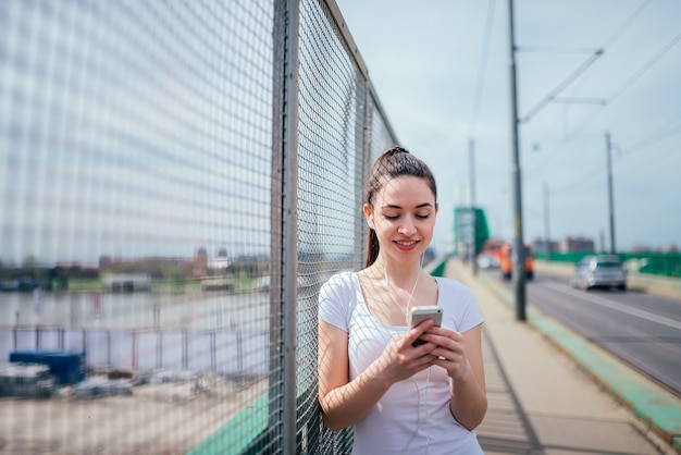 Smling girl leaning on wire fence. Listening music and using phone while standing on sidewalk.