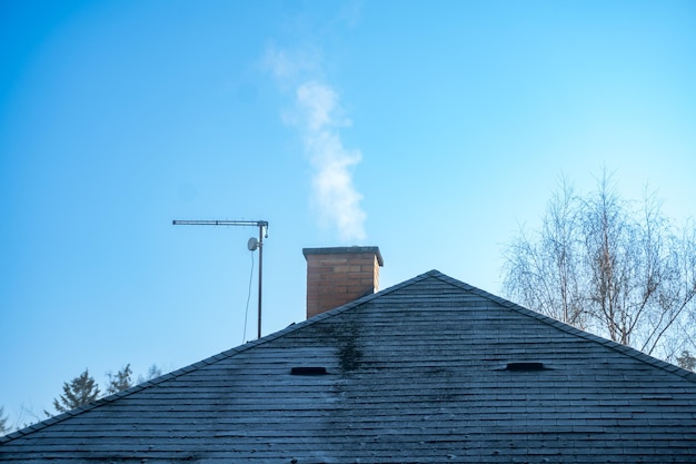 Smoke rises from the chimney on the house roof with smoking chimney and trees