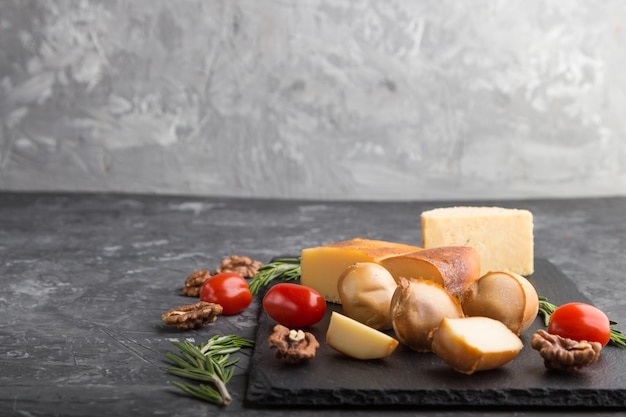 Smoked cheese and various types of cheese with rosemary and tomatoes on black slate board on a black concrete background. Side view, copy space