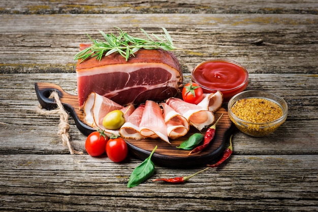 Smoked ham on wooden table with rosemary, pepper and cherry tomatoes.