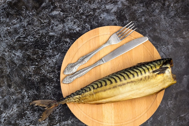 Smoked mackerel without head with fork knife cutting board on black background