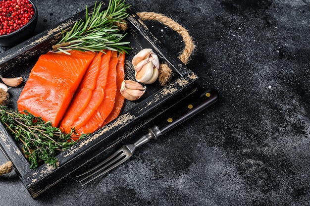 Smoked sliced salmon fillet in a wooden tray with herbs. Black background. Top view. Copy space.