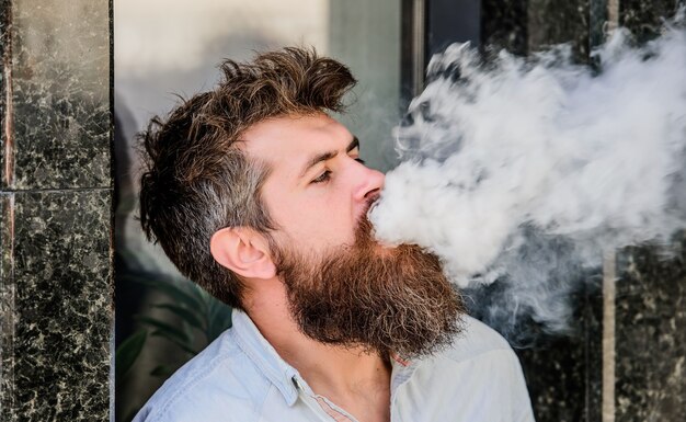 Smoking electronic cigarette. Stress relief concept. Smoking device. Man long beard relaxed with smoking habit. Clouds of flavored smoke. Bearded man smoking vape. Man with beard breathe out smoke.