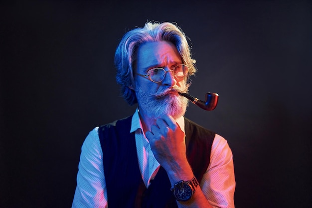 Smoking and having fun Neon lighting Stylish modern senior man with gray hair and beard is indoors