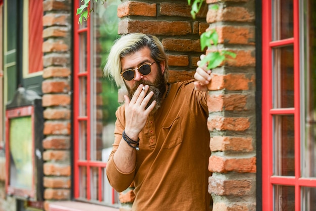 屋外での喫煙煙の休憩に行きました喫煙習慣タバコを持ったファッショナブルな成熟した男残忍な男サングラス喫煙タバコクールな男リラックスヒップスター喫煙古い建築の背景