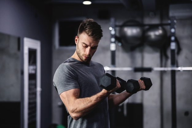 The smoldering look of an attractive and muscular man. A man in sportswear pumps his arm muscles and lifts dumbbells in a modern gym. Close up shot of a male person