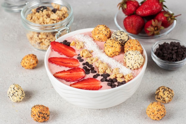 Smoothiekom met aardbeien, muesli, chocolade, kokosvlokken en knapperige honingballen in witte kom op grijze achtergrond