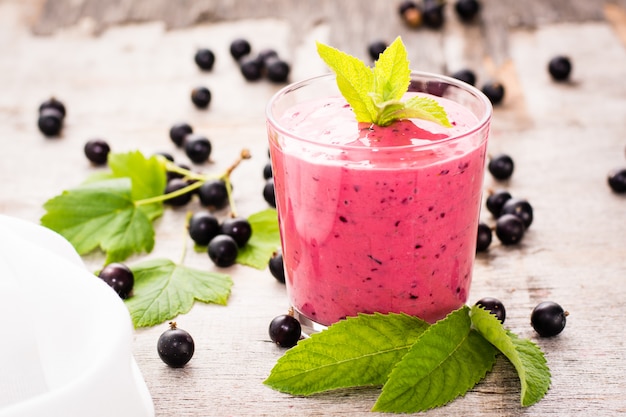 Smoothies with black currant and yogurt and mint leaves. Transparent glass standing on a wooden table surrounded by berries and leaves