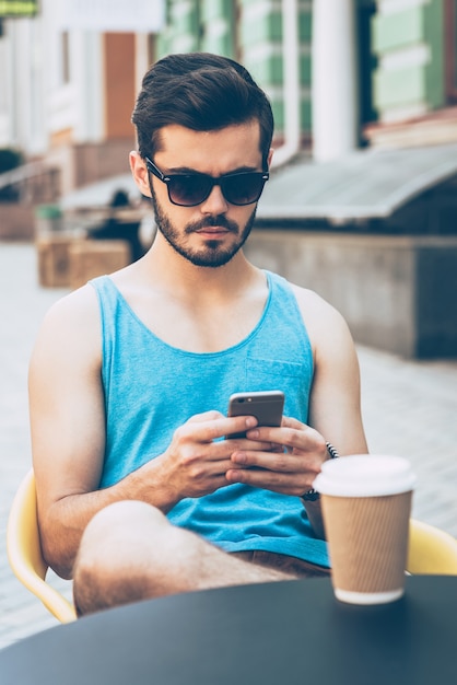 Sms'en naar vriend. Knappe jongeman in vrijetijdskleding die een smartphone vasthoudt en ernaar kijkt terwijl hij buiten in een café op de stoep zit