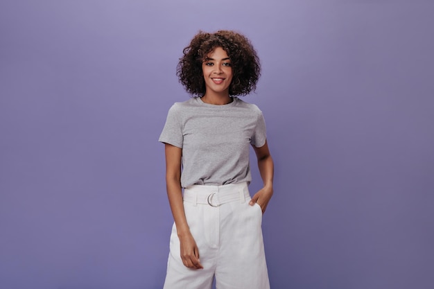 Snapshot of curly woman in white shorts against purple background Charming brunette girl in grey tee smiling on isolated backdrop