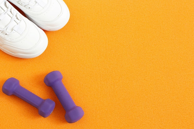Кроссовки и гантели на фоне оранжевого коврика для фитнеса. Плоская композиция. Концепция спорта, фитнеса, здорового образа жизни. Вид сверху. Скопируйте пространство.