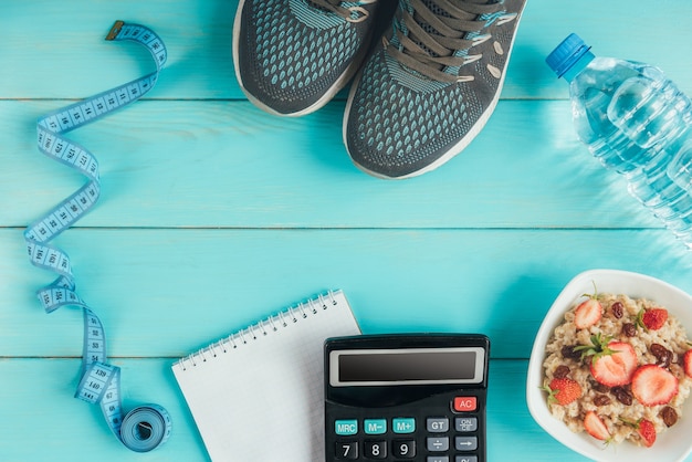 Sneakers, tape measure, notebook, calculator, bottle of water, apple and oatmeal with strawberry and raisins on blue, flat lay