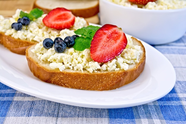 Sneetjes brood met wrongelroom, munt, bosbessen en aardbeien, een kom kwark, brood op een bord op een achtergrond van het blauw geruite tafelkleed