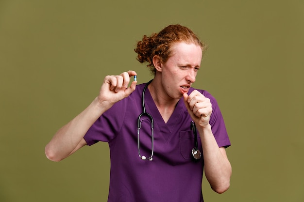 Sneeze holding pills young male doctor wearing uniform with stethoscope isolated on green background