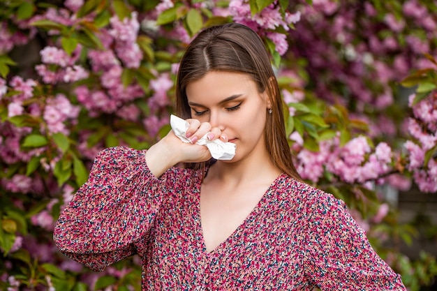 Sneezing young girl with nose wiper among blooming trees in park pollen allergy girl sneezing allergy sneezing spring woman sneezing in front of blooming tree spring allergy concept