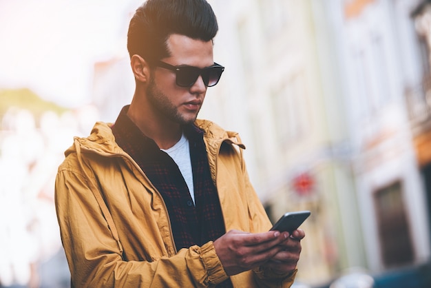 Snel bericht aan vriend. Knappe jonge man die een smartphone vasthoudt en ernaar kijkt terwijl hij op straat staat