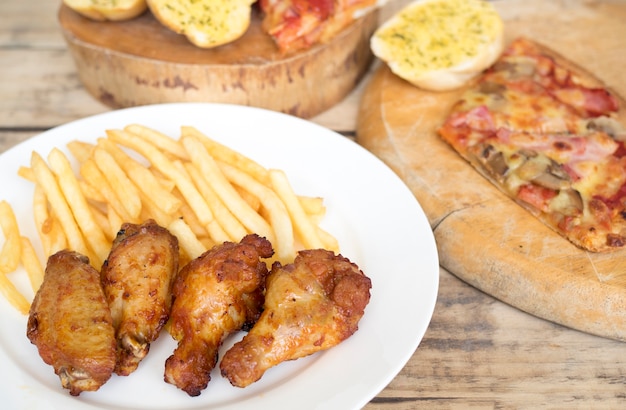 Snel voedsel, knapperige kippenvleugels, brood, frieten en pizza op houten achtergrond