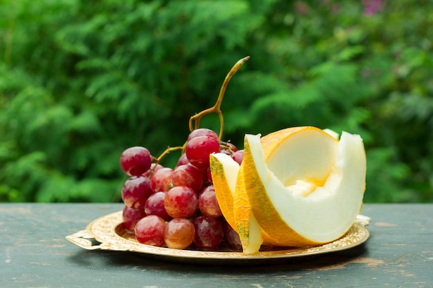 Snijd plakjes rijpe gele meloen en een tros druiven op een tafel met een natuurlijke groene achtergrond.