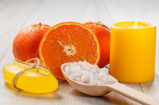 Snijd sinaasappel met twee hele sinaasappels, zeep, houten lepel met wit zeezout en brandende gele kaars op houten bureau. Spa-producten en accessoires
