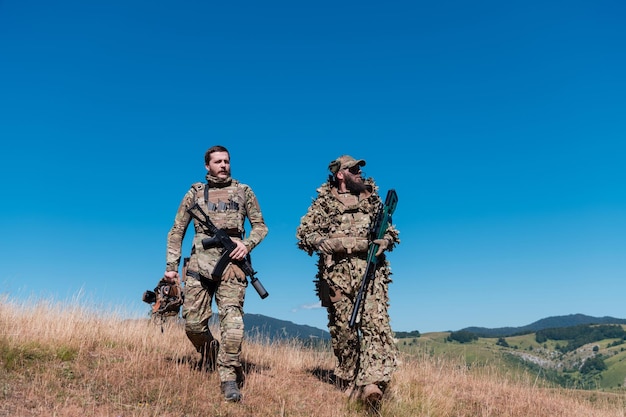 A sniper team squad of soldiers is going undercover. Sniper assistant and team leader walking and aiming in nature with yellow grass and blue sky. Tactical camouflage uniform.