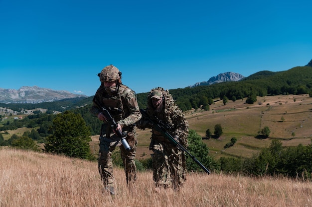 Под прикрытием действует снайперский отряд солдат. Помощник снайпера и руководитель группы гуляют и целятся на природе с желтой травой и голубым небом. Тактическая камуфляжная форма.